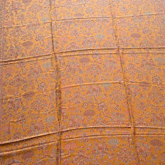 A rectangular woven textile laid flat on a light gray floor, featuring an intricate floral pattern in orange, gold, and muted blue, with striped multicolor borders along the edges.