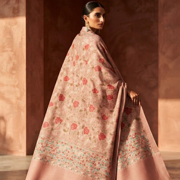A woman wearing a flowing dusty pink shawl with red and green floral motifs and a wide border, photographed in a warm-toned architectural setting.