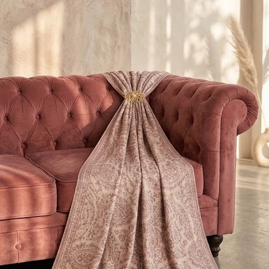 A draped dusty rose pink patterned throw blanket with gold fringe and a central gold tie, styled on a matching pink tufted sofa in a sunlit room.