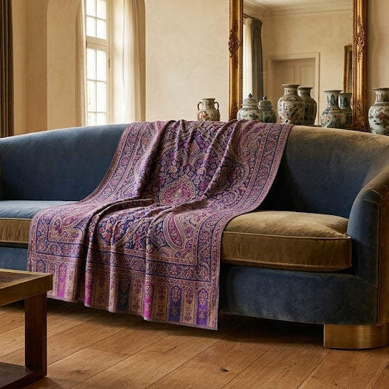 A curved velvet sofa in deep blue and tan upholstery, styled with a purple patterned shawl, set in a warm, classic room with wood floors, a wooden coffee table, and a bookshelf.