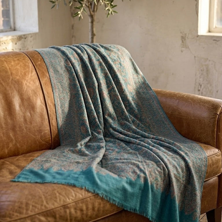 A teal and brown patterned throw blanket with fringed edges draped over a brown leather sofa in a rustic room setting.