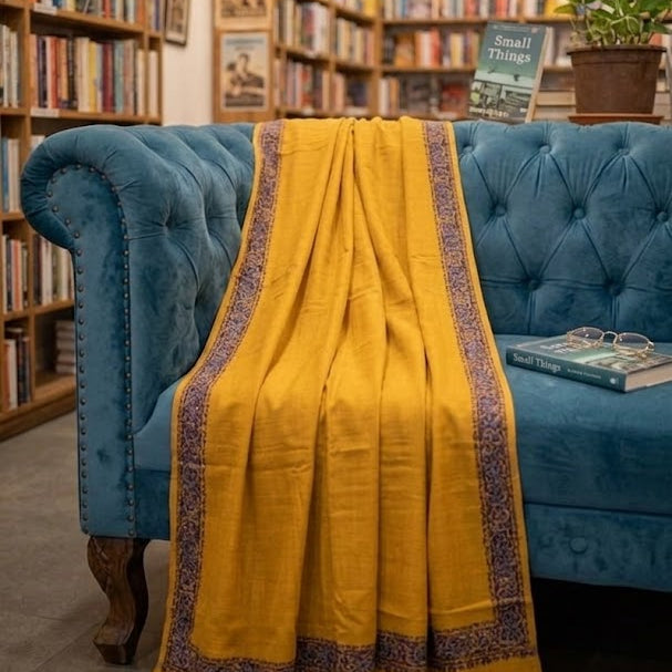 A mustard yellow shawl with a purple patterned border draped over a blue velvet armchair in a cozy, book-filled library setting.