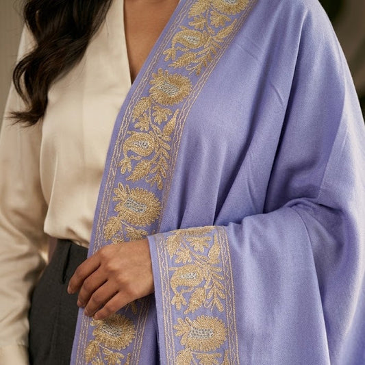 A close-up of a person wearing a lavender blue shawl with ornate gold floral embroidery along the borders, paired with a cream blouse and dark trousers.