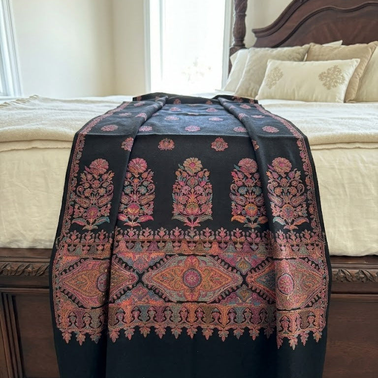 A black woven throw blanket with intricate pink, red, and gold floral and geometric patterns, draped over the foot of a bed in a bedroom setting.