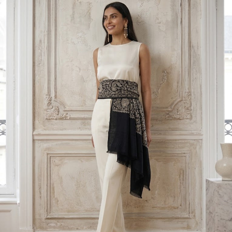 A woman models a sleeveless cream-colored jumpsuit with a wide black and beige patterned waist sash and a matching black draped scarf, standing in a classical interior with ornate wall paneling.