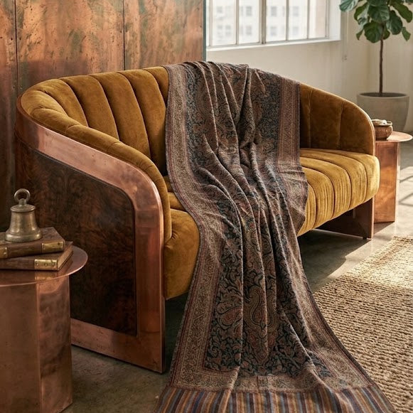 A styled interior scene featuring a curved velvet sofa upholstered in mustard yellow fabric with a dark brown patterned throw blanket draped over it. The sofa is set in a room with a large copper-paneled wall, a window, a potted plant, and a woven rug.