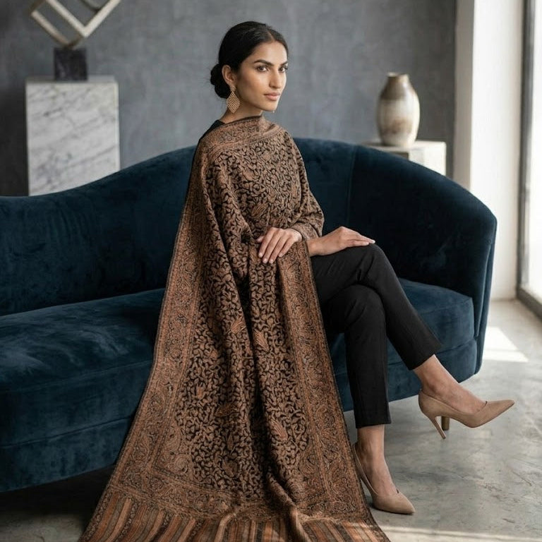 A woman sits on a dark blue velvet sofa wearing a brown patterned shawl draped over her shoulders, paired with black pants and beige heels in a modern room with a gray wall.