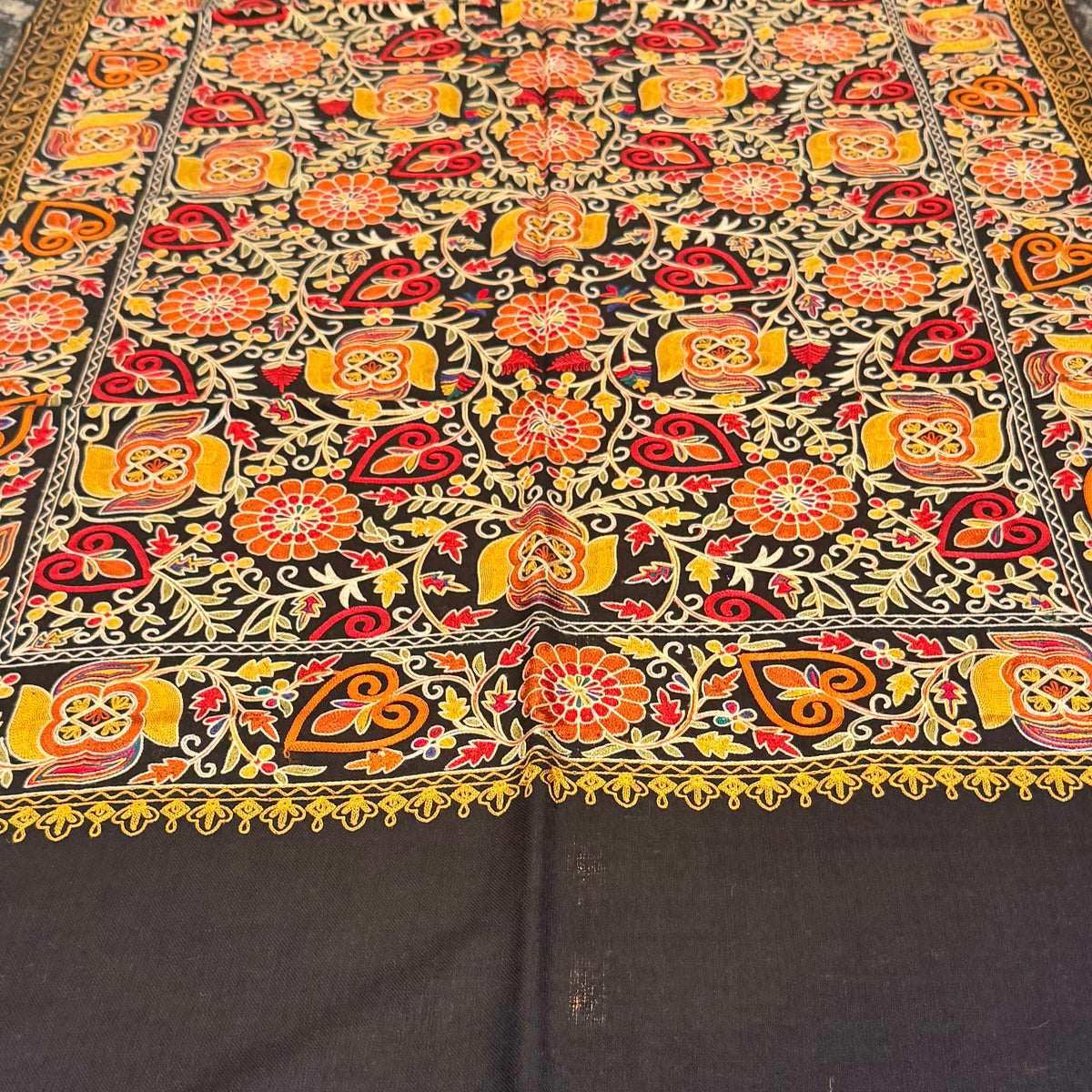 A rectangular table runner with a vibrant, intricate floral and geometric pattern in orange, red, yellow, and green, laid out on a dark marble dining table with chairs visible on the right side.
