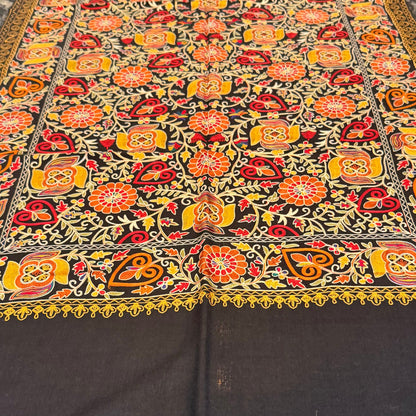 A rectangular table runner with a vibrant, intricate floral and geometric pattern in orange, red, yellow, and green, laid out on a dark marble dining table with chairs visible on the right side.