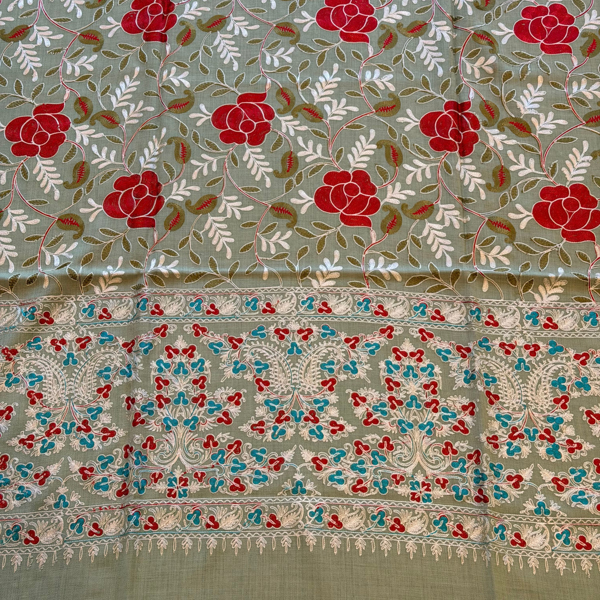 A close-up of a patterned fabric panel with a sage green base, featuring red floral motifs, white leafy vines, and a decorative border with red, blue, and white embroidery.