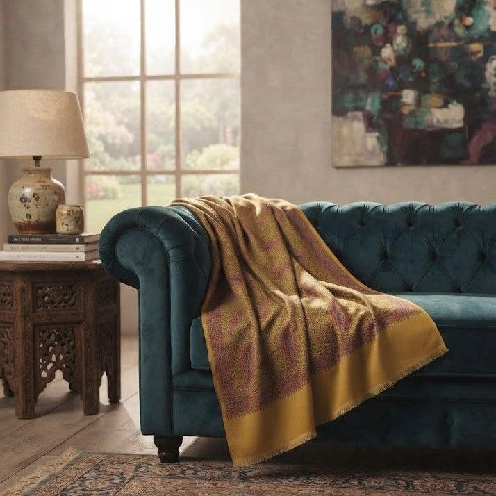A teal velvet tufted sofa in a styled living room, with a mustard yellow and burgundy patterned throw blanket draped over the arm. A wooden side table with a lamp, a large arched window, and a patterned rug are visible.