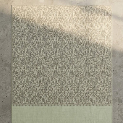 A rectangular woven sage green embroidered shawl, featuring an intricate beige damask pattern on the upper portion and a solid sage green border at the bottom. It is laid flat on a textured gray concrete surface with soft natural shadows.