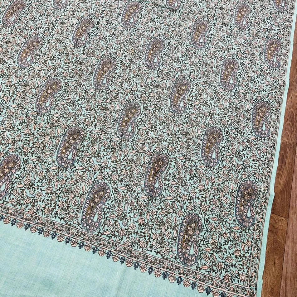 A light sage green silk Shawl with an intricate beige and brown paisley pattern, displayed flat on a wooden floor.