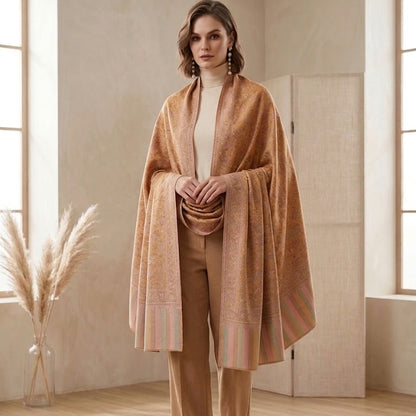 A woman stands in a softly lit room wearing a warm camel-colored shawl draped over a cream turtleneck, matching tan trousers, and black pointed heels. The room has light wood floors, beige walls, and a vase of dried pampas grass near the window.