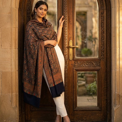 A woman stands in front of an ornate carved wooden door wearing a dark brown and rust patterned shawl over a white outfit. The shawl features a deep navy blue border.