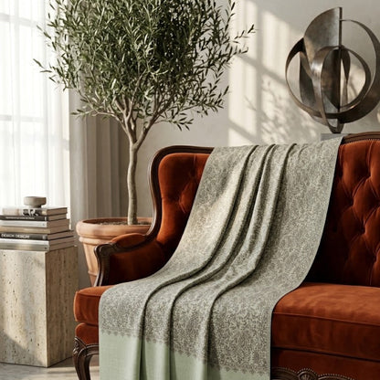 A sage green and gray patterned throw blanket with a solid sage green border is draped over a rust-orange velvet tufted armchair in a styled living room setting.