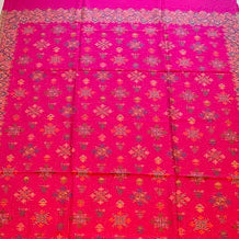 A bright red and magenta patterned shawl or scarf laid flat on a light wood floor, featuring intricate geometric and floral motifs in gold, orange, and blue, with a matching fringed edge.