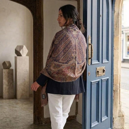 A woman wearing a patterned paisley shawl in brown, beige, and blue tones, a dark navy tunic, and white trousers, standing in a doorway next to a blue wooden door.