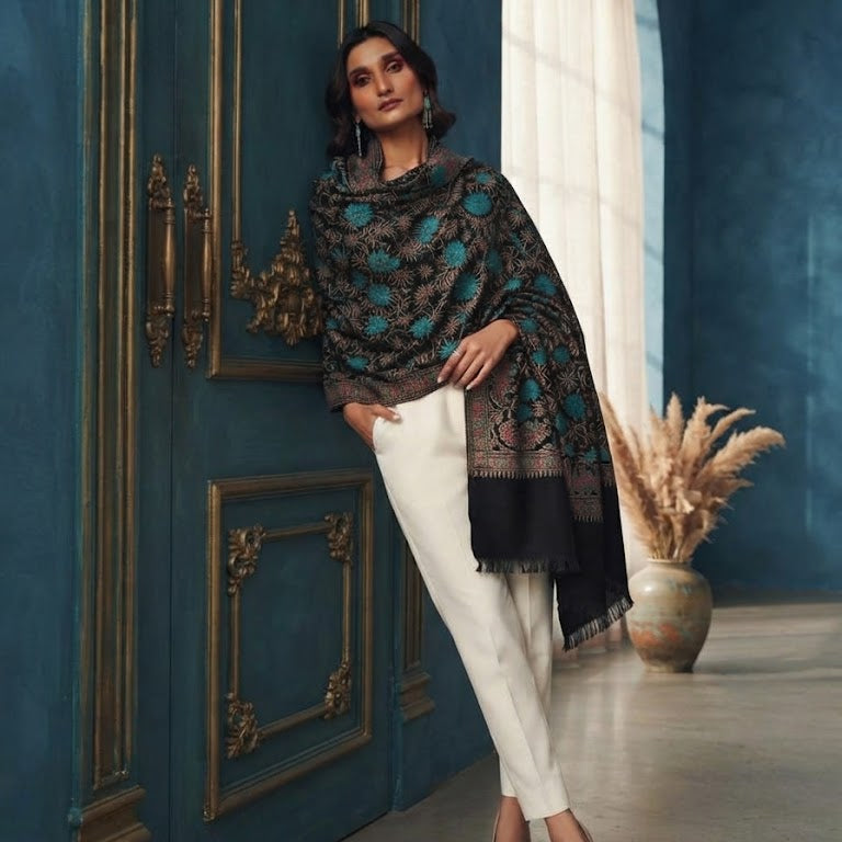 A woman models a black and teal patterned shawl draped over her shoulders, paired with white trousers and beige heels, standing in front of an ornate dark blue door.