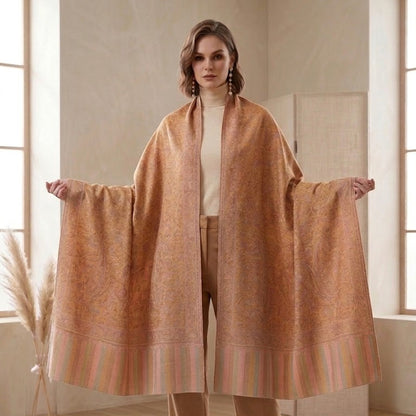 A woman modeling a large, open-front shawl or wrap in a warm camel-brown color with subtle woven patterns and striped borders, styled with beige trousers and black heels in a bright, modern room.
