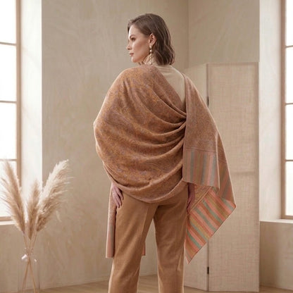 A woman stands in a softly lit room wearing a large draped camel-brown shawl over her shoulders and tan trousers, shown from the back. The room has light wood floors, beige walls, and a vase of pampas grass near a window.