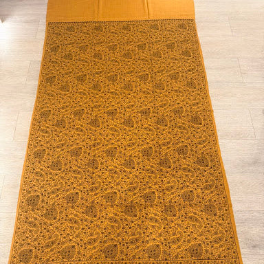 A mustard yellow saree with an intricate brown paisley and floral pattern, laid flat on a light gray tiled floor.