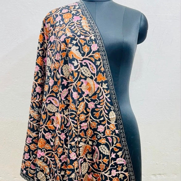 A black embroidered shawl or dupatta with intricate floral and paisley motifs in orange, pink, and green, draped over a black mannequin torso against a light gray wall.