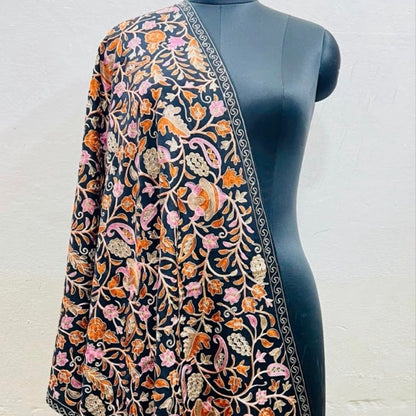A black embroidered shawl or dupatta with intricate floral and paisley motifs in orange, pink, and green, draped over a black mannequin torso against a light gray wall.