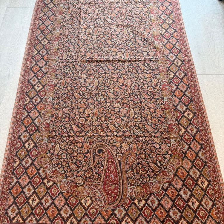 A traditional Persian-style area rug with an intricate floral and paisley pattern, laid out on a light-colored wooden floor. The rug features a dominant dark brown center field surrounded by a red and black diamond-patterned border with beige and red accents.