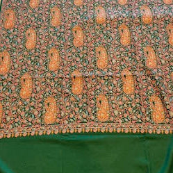 A green and brown patterned shawl , likely a saree or scarf, laid flat on a light wood floor. The fabric features a solid dark green border at the top and bottom with a central patterned section containing brown paisley motifs and intricate floral details on a beige background.