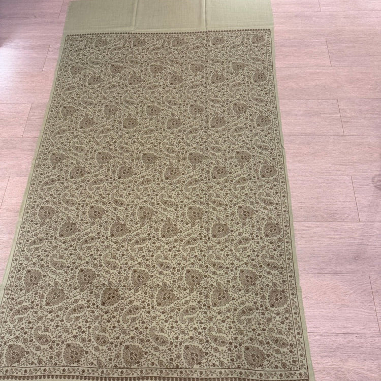 An olive green woven runner or scarf with an intricate beige and brown paisley pattern, laid flat on a light-colored wooden floor.
