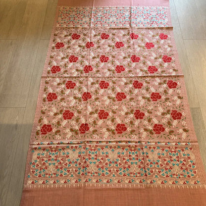 A peach-colored woven textile, likely a dupatta or sari, featuring red floral motifs and intricate multicolor embroidery, laid flat on a light wood floor.