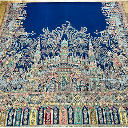 A large traditional oriental rug with an intricate architectural and floral pattern, featuring a deep blue field, cream and gold central design, and a multicolored border, laid out on a light wood floor.