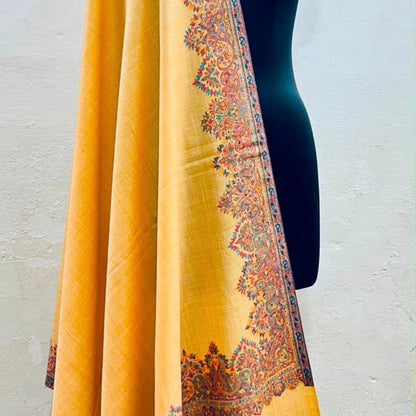 A mustard yellow shawl with an ornate multicolor paisley border, draped over a black mannequin torso against a light gray wall.