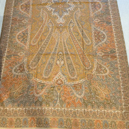 A large vintage-style oriental rug with an intricate medallion and paisley pattern, laid flat on a light-colored wooden floor. The rug features a dominant ochre-yellow center with rust orange, beige, and muted gray-blue details.