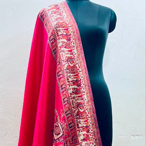 A bright pink and red dupatta with an intricate multicolor traditional border pattern, draped over a black mannequin torso against a white wall.