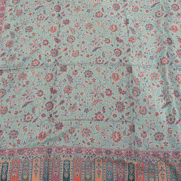 A long rectangular patterned textile laid flat on a light wood floor, with a pale blue-green base and dense floral motifs in pink, red, and teal, plus decorative striped borders at both ends.