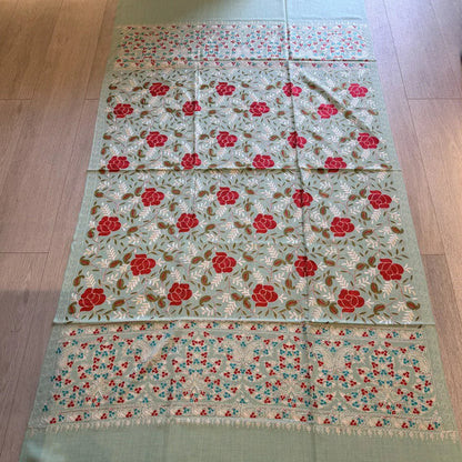 A long mint green embroidered textile, likely a table runner or decorative mat, laid flat on a light wood floor. The fabric features red floral motifs and green leafy embroidery throughout, with a solid mint green border at the bottom.