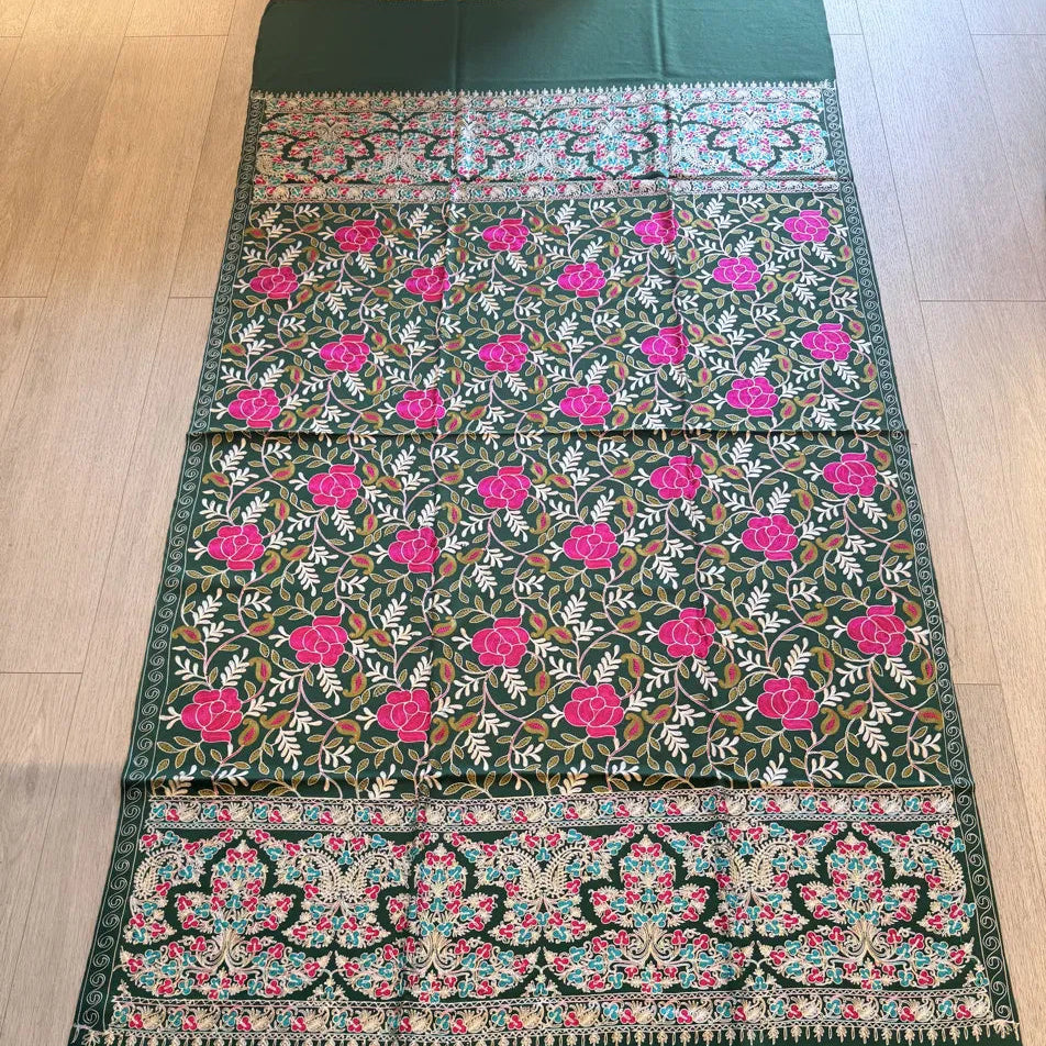 A dark green embroidered fabric, likely a saree or scarf, laid flat on a light wood floor. The fabric features vibrant pink floral motifs, green and white leafy patterns, and ornate borders with pink, green, and white embroidery.
