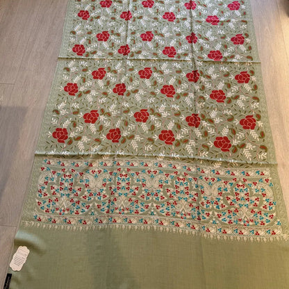 A light sage green embroidered fabric, likely a shawl or scarf, laid flat on a light wood floor. The fabric features red and white floral motifs with green and gold embroidery, and a decorative border with red, blue, and white floral details.