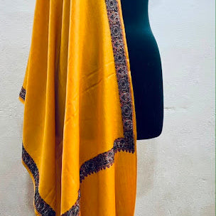 A mustard yellow shawl with a dark navy blue patterned border, draped over a black dress form mannequin against a white wall.