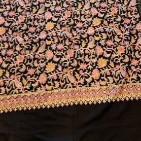 A black woven textile, likely a Zari scarf or sari, featuring an intricate floral pattern in pink, orange, and yellow, laid flat on a light wood floor.