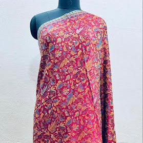 A long-sleeved, one-shoulder traditional dress displayed on a black mannequin torso against a plain off-white wall. The garment features an intricate, dense floral and paisley block print in shades of red, pink, orange, and blue.