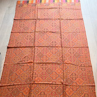 A long patterned textile laid flat on a light wood floor, featuring an orange and red floral motif with multicolored striped borders at both ends.