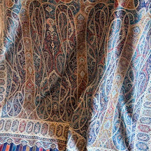 A large patterned throw blanket draped over a chair in a home setting, featuring an intricate paisley design in shades of blue, red, beige, and brown.