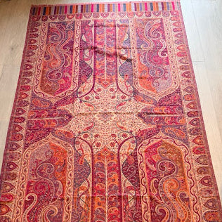 A large rectangular patterned rug laid flat on a light wood floor, featuring intricate paisley and floral motifs in red, pink, orange, beige, and purple tones.