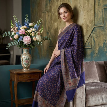 A woman wearing a purple patterned kani jamawar shawl with a beige border sits on a gray velvet sofa in a decorated room with a wooden side table and a floral arrangement.