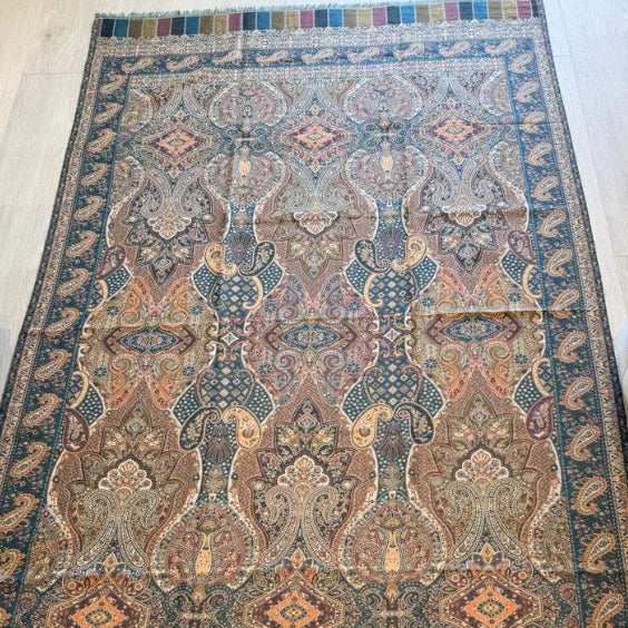 A rectangular traditional area rug with an intricate geometric and floral pattern in muted blue, beige, brown, and rust tones, laid out on a light wood floor between a sofa and a bench.