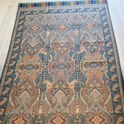 A rectangular traditional area rug with an intricate geometric and floral pattern in muted blue, beige, brown, and rust tones, laid out on a light wood floor between a sofa and a bench.