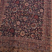 A long rectangular shawl with an intricate floral pattern in dark red, purple, and brown tones, laid out on a light wood floor next to a beige upholstered chair.